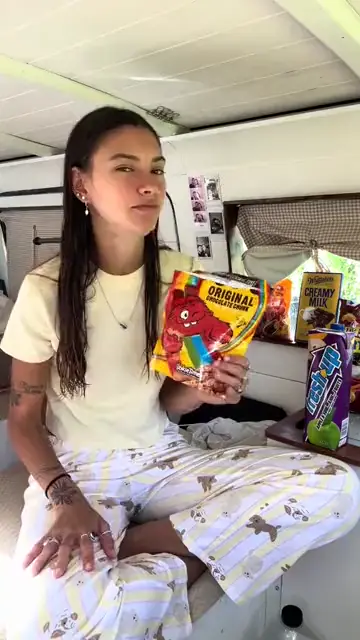 A woman sitting cross-legged holding up a bag of chips to the camera